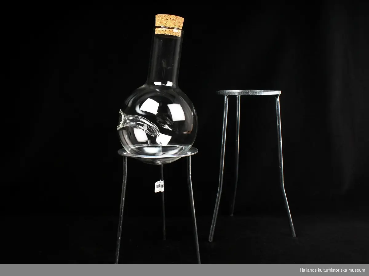 A. Flaska av glas, glasskulptur med två tillhörande stativ (B och C) av vitmetall. Helt av transperant glas med tillhörande korkplugg.
Tungliknande infällning i glaset.