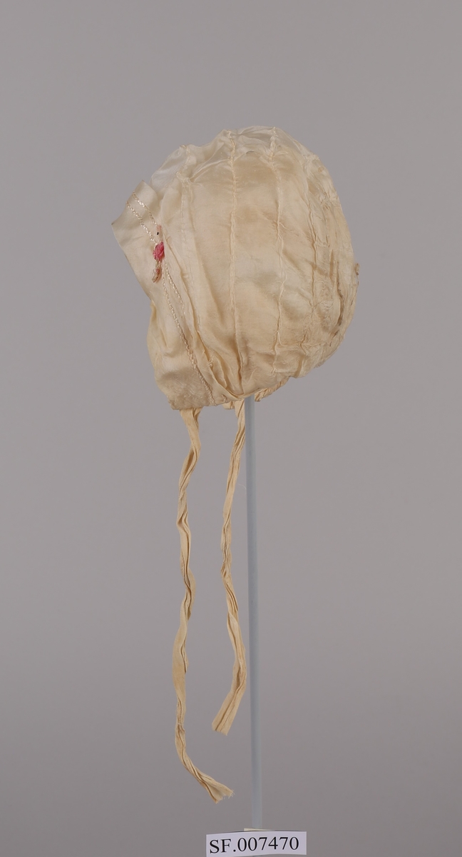 Dåpshue i silke med to band for knyting under haka. Brodert litt bak nederst på venstre side, ein rosa blome.