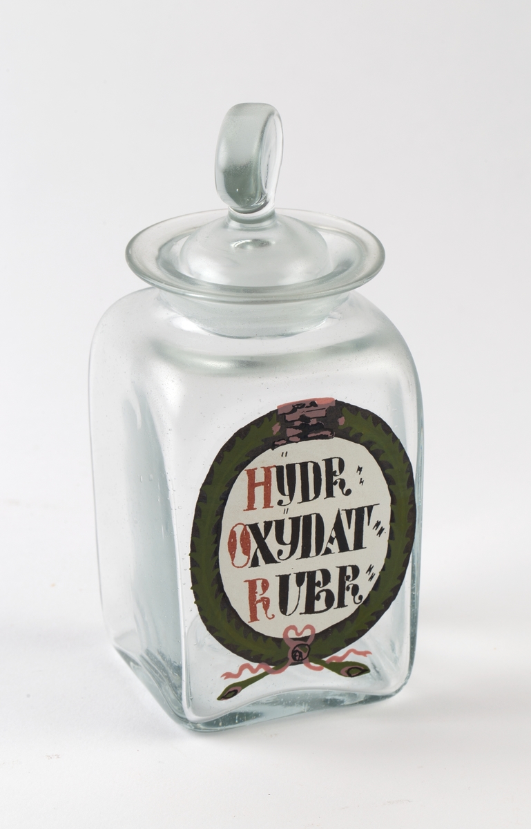 Kubeformet glassbeholder med bred sirkulær åpning og glasspropp. Oval påmalt etikett med sort og rød skrift, pyntet med grønn bladkrans og rosa sløyfe.