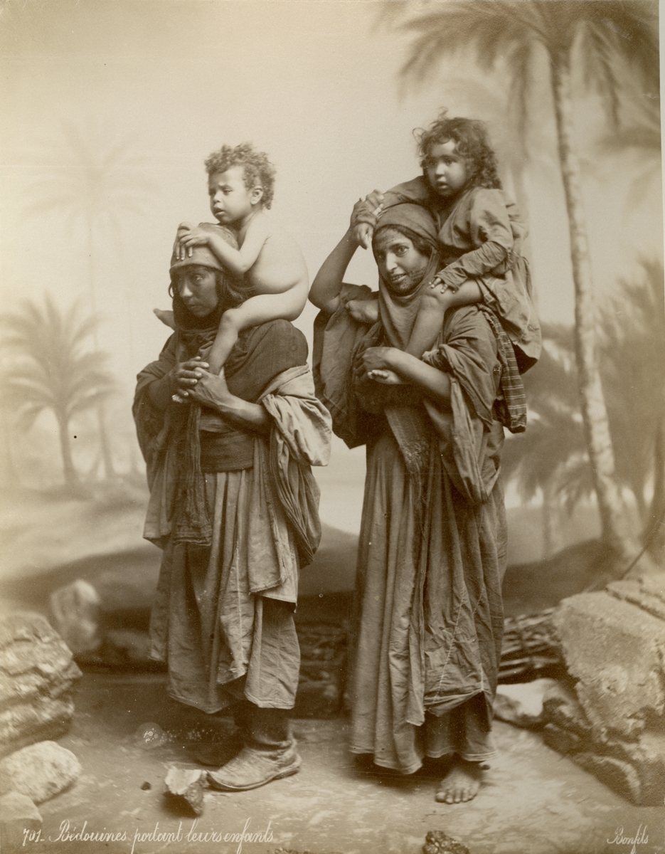 Porträtt av två kvinnor bärande två små barn. Ateljebild med bakgrund, kvinnorna är eller ska föreställa beduiner.