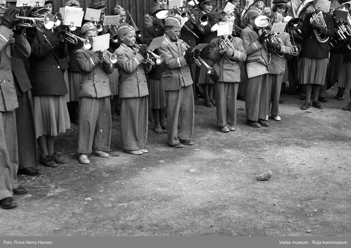 Vadsø sentrum 17. mai 1951. Deler av flere musikkorps i forskjellige uniformer, bestående av barn og voksne står oppstilt og spiller. Korpset står foran postbrakka.