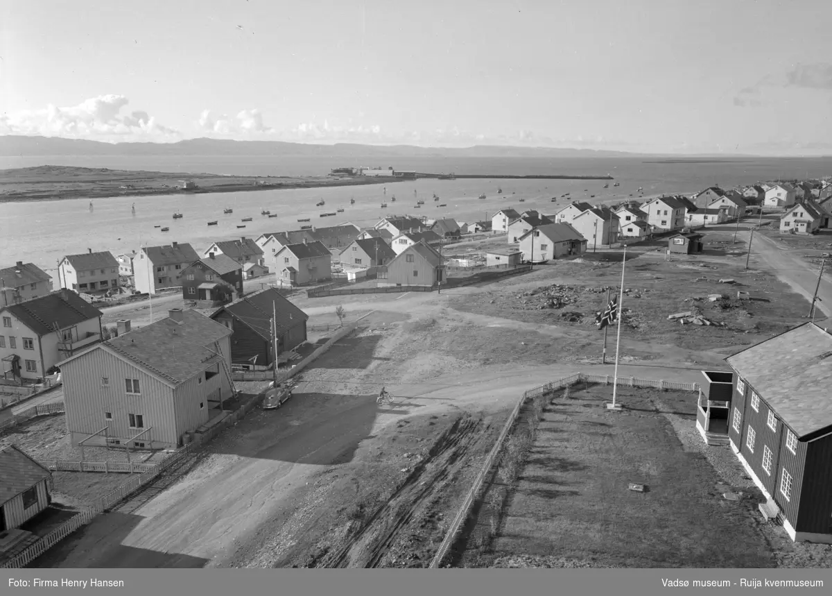 Vadsø 1957. Bildet er tatt fra kirken (som sto ferdig i 1958). Bildet viser boliger langs Damsveien og Nyborgveien. Til høyre og nærmest kamera vises en del av presteboligen. På havna ligger mange båter. Vi ser en del av øya med Vadsø sildoljefabrikk og moloen.