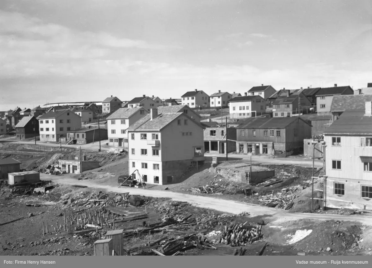 Vadsø 3.6.1951, sentrum. Bildet er tatt fra sørøst mot nordvest, trolig fra et hustak sør for torget. Bildet viser bygninger nord for Kaigata og langs Havnegata, både ferdige og uferdige bygg og brakker. I forkant en del av fjæra sør for Kaigata.
