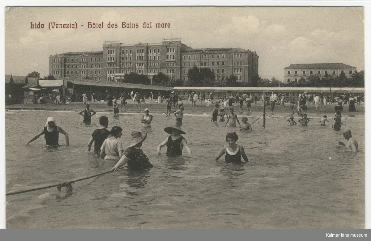 Badande män och kvinnor i mer eller mindre heltäckande badkläder, några bär bredbrättade hattar. I bakgrunden stranden och stora byggnader. Grand Hotel des Bains byggdes 1900.