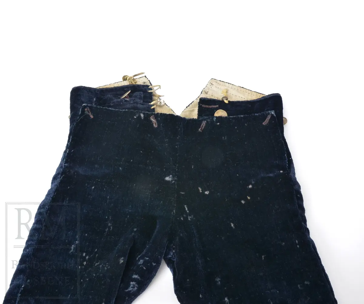 Mørk blå fløyelsbukse, knelang, med lem. Foret i lin. Fem flate messingknapper.