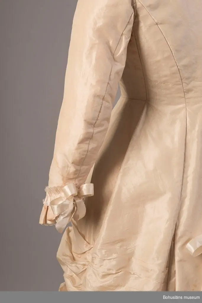 Gulvit klänning buren av Pauline Ekelund, född Berg (1843 - 1881), vid giftermål 1879 med överstelöjtnant Frithiof Ekelund. Fotografi från bröllopet, se bildarkivet UMFA samt ramat färglagt porträtt UM018507.
Inprovat liv, avskuren i sidorna, öppen fram, knäpps med tygkädda knappar. Halskant garnerad med gulvit, veckad bomullsspets, maskintillverkad samt kant av veckat siden med uppfransade kanter. Lång ärm avslutad med volang av tyget. För övrigt garnerad på samma sätt som kring halsen. Rosett av gulvitt sidenband fastsytt vid handleden. Lång kjol med släp. Rynkad mitt fram och vid sidorna. Baktill är kjolen skuren för turnyr. Släp garnerat med tre rosetter av gulvitt sidenband. Kjolkanten garnerad på ovansidan med veckat, tunt bomullstyg samt veckad remsa av tyget med uppfransade kanter. Under kjolfållen veckat bomullstyg som på ovansidan, kantad med smal, veckad kant, kantad med vit spets. Kjolfållen skodd med stärkt, vitt bomullstyg. Livet fodrat med vitt bomullstyg. 

Se bilagepärm med kopia av "inbjudan till vigselakten mellan Kaptenen vid Kongl. Bohus Läns Regemente Frithiof Ekelund och Pauline (född Berg) tisdagen den 7 oktober 1879 i Rådhussalen, Uddevalla". 

Ur punktnummerkatalogen 1957-1978:
Fru Anna Barks, sterbhus