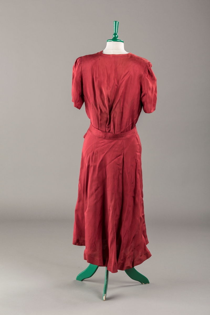 Form: Glatt, klokkeformet skjørt. Forstykkene rynket til buede bærestykker foran. Overdel rynket til skjørt. Korte ermer. Lukket med trykknapper. Belte fastsydd.