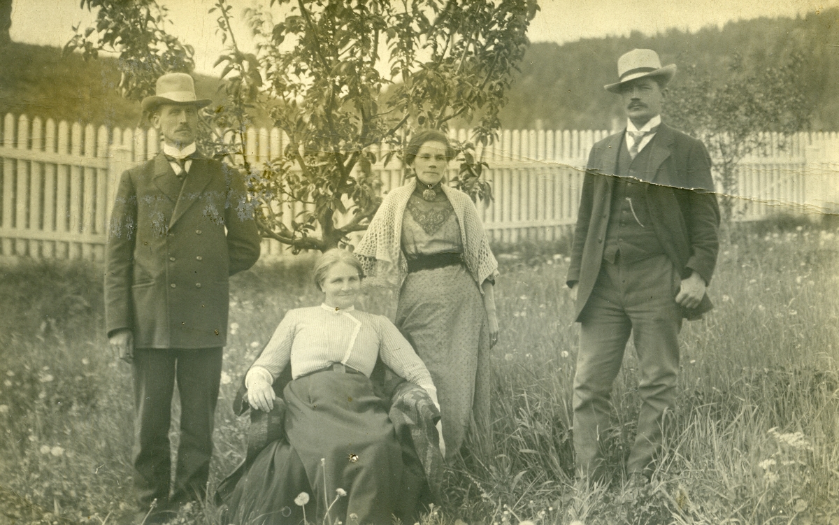 Postkort. Fire personer avbildet i en hage. 2 kvinner og 2 menn. Epletre og stakittgjerde i bakgrunnen.