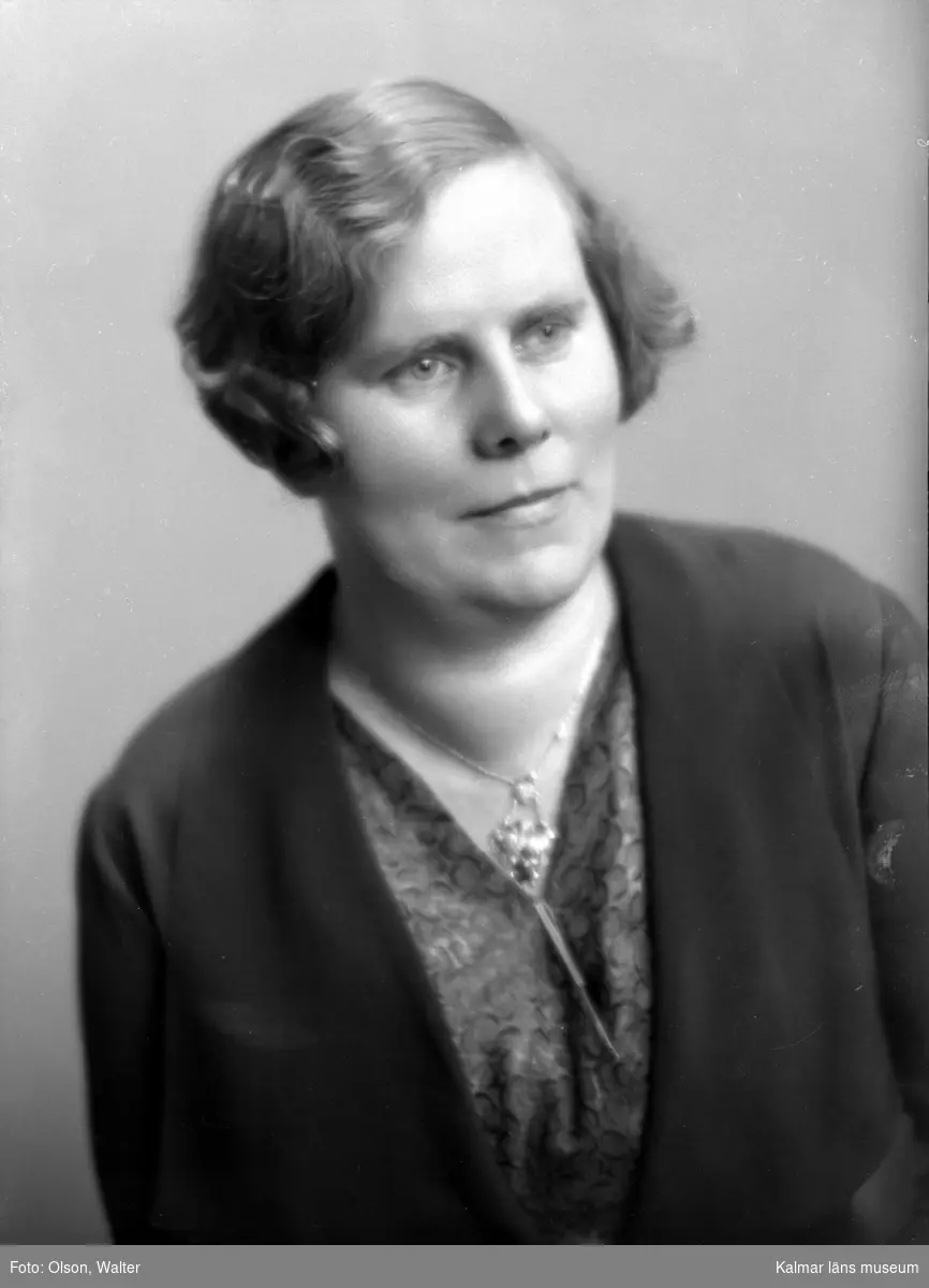 Ateljébild på en kvinna i halsband och jacka. Enligt Walter Olsons journal är bilden beställd av fru Helga Nilsson.