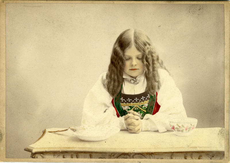 Kolorert studiofotografi av jente med Hardangerdrakt, sitter med foldelde hender på bord. 1903. (Foto/Photo)