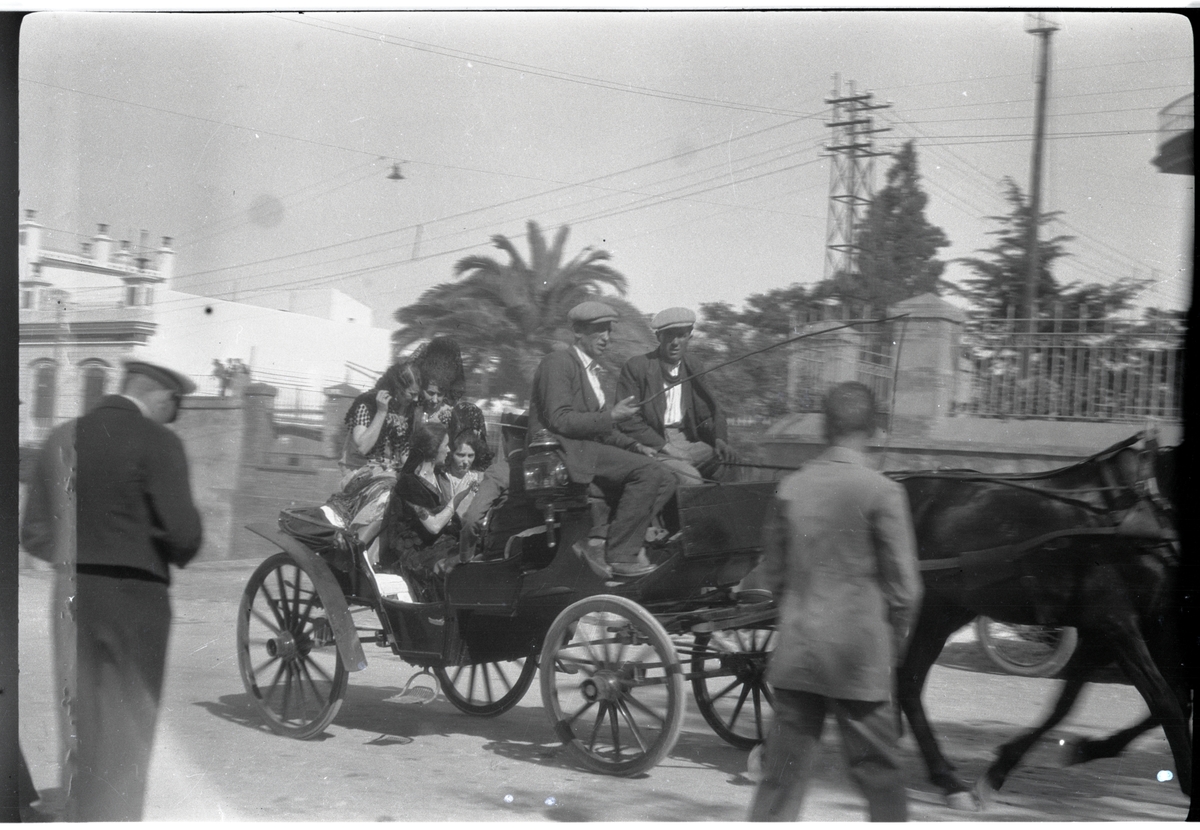 Häst och vagn med två män på kuskbocken samt fyra kvinnor och en man som passagerare. I v hörn står en man, som förefaller bära svensk officersuniform och titta ner i ljusschaktssökaren på en kamera.