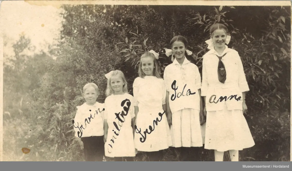 amatørfotografi av fire jenter og ein gut kledd i kvite klær som står på rad framfor busker