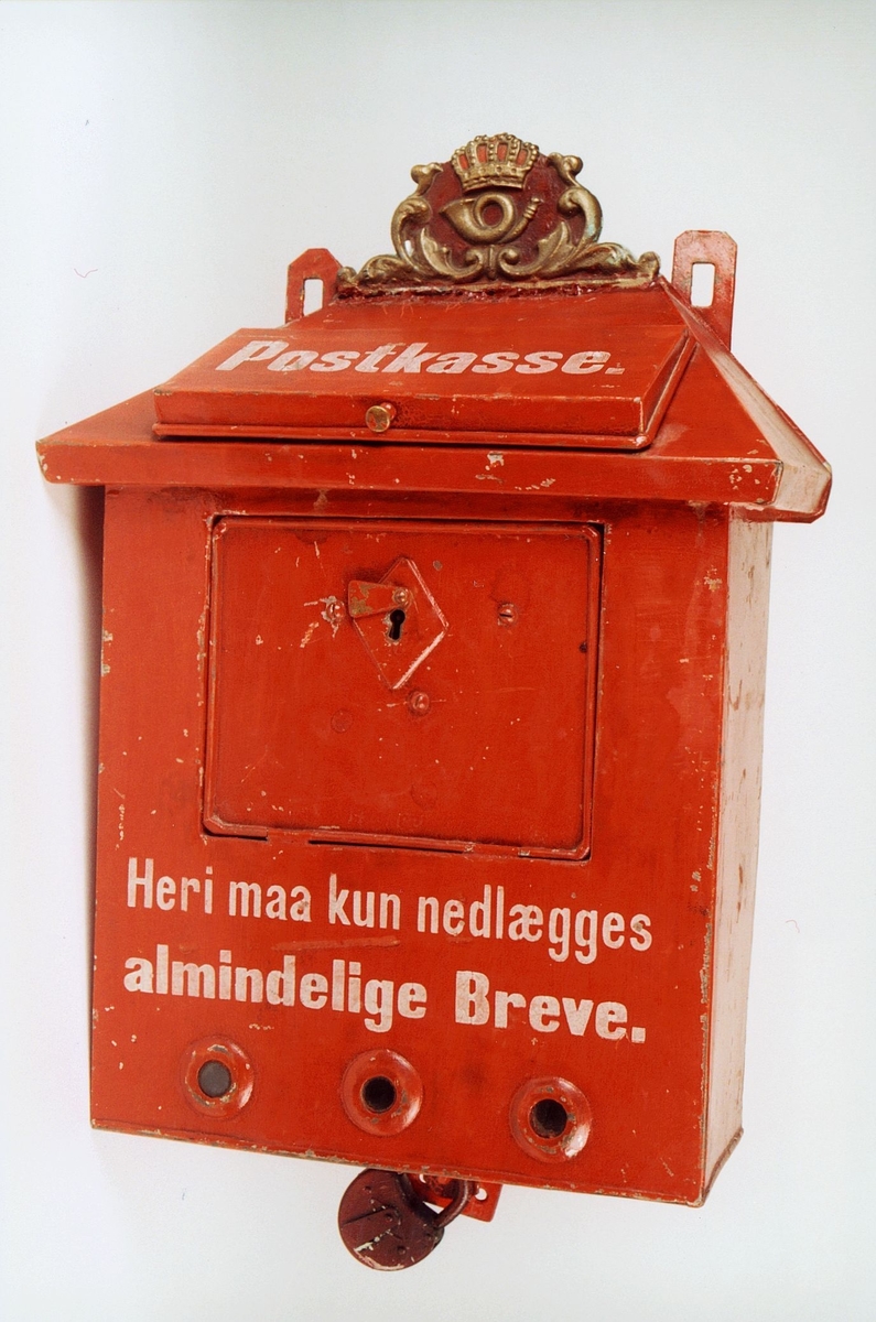 Rød postkasse utsmykket med posthorn og krone øverst.

Tekst på kassen: "Postkasse. Heri maa kun nedlægges almindelige Breve".