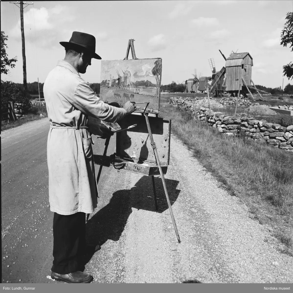 En konstnär i hatt och målarrock står vid ett staffli på en grusväg och målar av kvarnar bakom en låg stenmur.