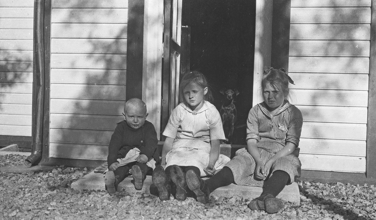 Gruppebilde. Tre barn, en gutt og to piker som sitter på en trapp i solskinnet. En dackshund står bak i døråpningen og ser ut. Hverdagsfoto.