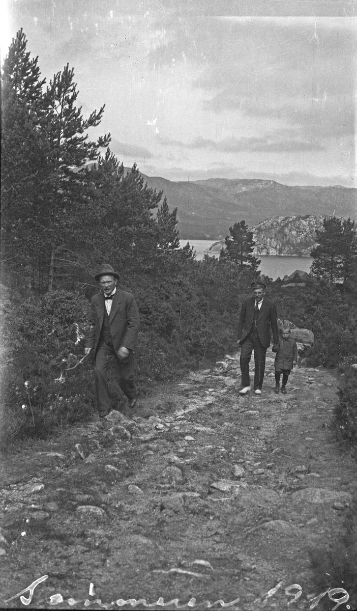 Sommer. To menn på vei opp en lang bakke på en skogsvei. En liten gutt med kortbukser er med. Fjorden i bakgrunnen. Hverdagsfoto.