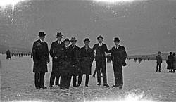 Vinter. Syv menn i hatt og frakk poserer på isen. De fleste 