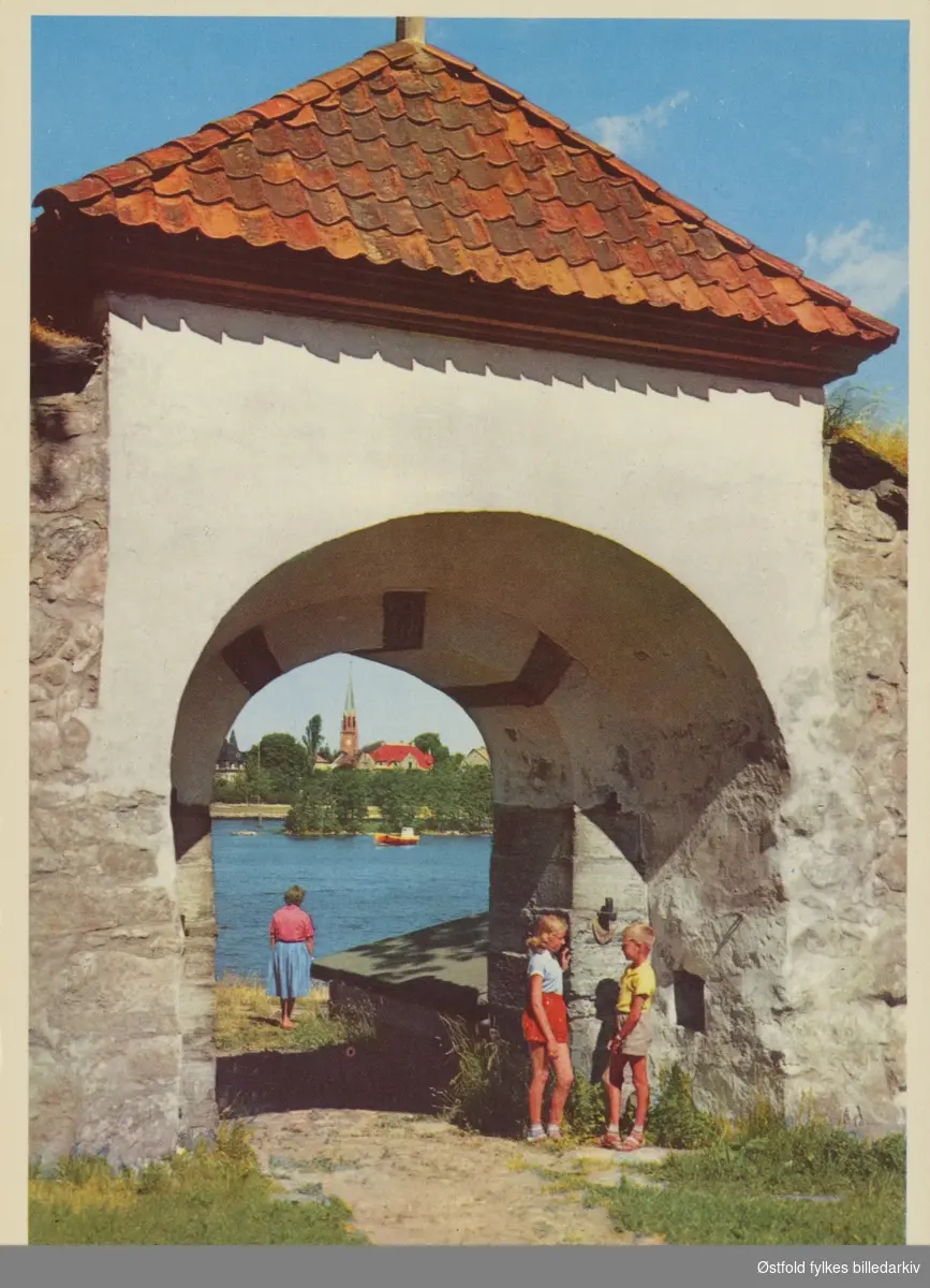 Mellomporten til Gamlebyen i Fredrikstad, med Glomma og Vestsiden kirke i bakgrunnen. Gutt og jente står i porten, ukjente. Postkort.