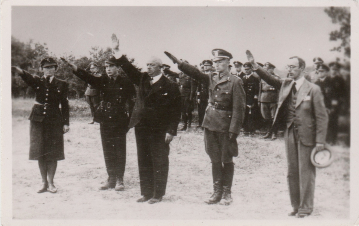 En kvinne og fire menn gjør nazihilsen. Tre av dem er uniformert, to i hirduniform og en i politiuniform modell 1942. En flokk menn i tyske uniformer skimtes i bakgrunnen.