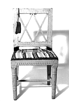 Vitmålad gustaviansk stol, svarvade framben med räfflor. Raka sarger med skuren dekor i form av nagelskärning, äggstav, pärlstav, i hörnen skuren stjärna inuti en cirkel. Kvadratisk genombruten rygg med snedställda spjälor i form av 2 kryss har ursprungligen haft  påpinnad sits, på senare tid omgjord till fast stoppning.