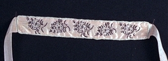 Brudstrumpeband av ljust rött siden med broderier av pärlor i svart och vitt samt silke i svart bestående av fem buketter.
Kantat med vit snodd av sammet, baksidan av vitt siden.