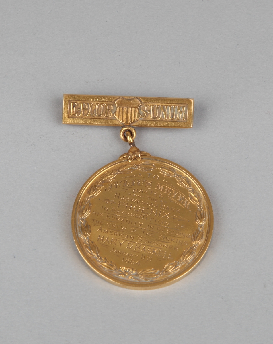 Rund gullmedalje med nål med motiv av kvinnehodeog tekst på forsiden samt tekst på nålfeste og baksiden.