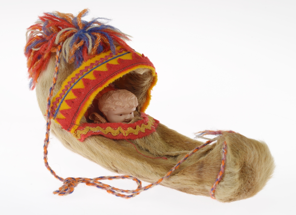 En liten komse med en pute og et dukkehode i. Gjenstandene er undernummerert 01-03. 
FTT.54491.01: En liten komse laget av et uthulet stykke tre som er kledd med garvet reinskinn. På kalesjen er det dekorert med røde pyntebånd av ull festet på et gult bånd av ull. Det er også to flerfargede dusker på kalesjen. i kalesjen er det festet et tvunnet ullbånd som er festet i den andre enden av komsen slik at man kan bære den. Det er også sydd på et pyntebånd i gult og rødt på oversiden.
FTT.54491.02: En liten rød pute sydd av silkestoff som ligger inni komsen.
FTT.54491.03: Et dukkehode laget av plast som ligger i komsen. Håret er støpt og er krøllete. I ansiktet er det malt på øyne og munn. Rundt halsen har dukken et silkebånd.