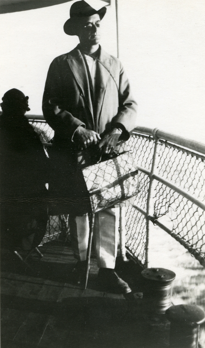 Mann, Haakon Garaasen (6/7 1887 - 1957), med hatt, stav, veske og briller, stående i sola på skipsdekk.