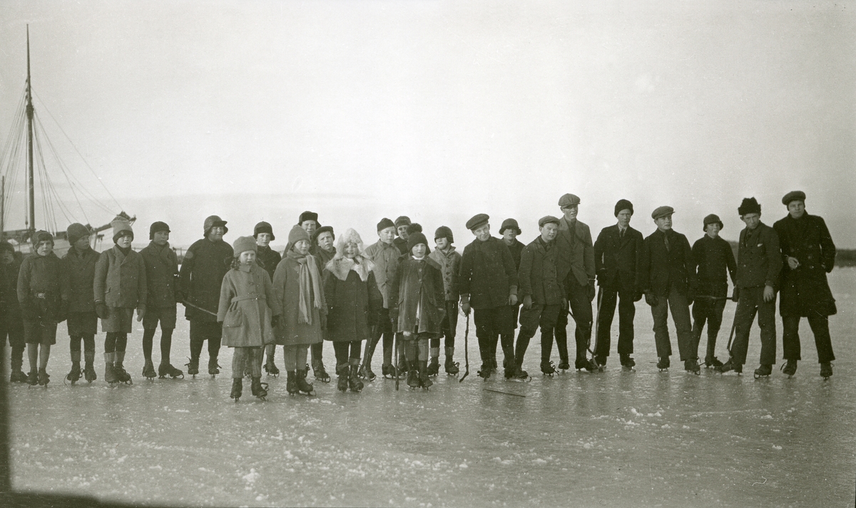 "Skokk" med unger stående på isen med skøyter på bena i Fredriksvern (Stavern). Fiskeskøyte (el. lign.) skimtes i bakgrunnen til venstre.