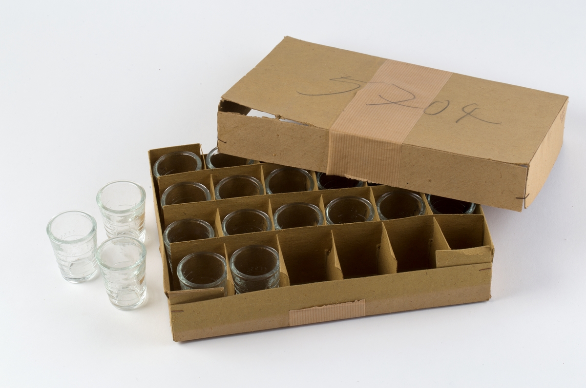 Eske av kartong med lokk. Inndelt i 24 rom med 23 måleglass. Hvert måleglass med innstøpt gradering på ene siden opp til 20 ml, på annen side 1 TE EL / 1/2 E SSL / 1 ES SL.
Esken er holdt sammen med hyssing. På lokket notert med blyant: 5704