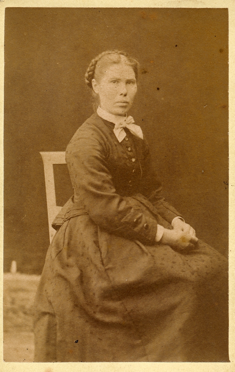 Portrett, ung kvinne som sitter på en stol med hendene i fanget. Oppsatt hår, flette.