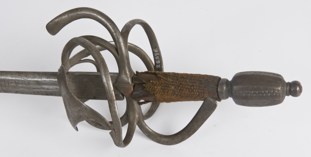 Plommeformet  8 kantet knapp. Tregrep.. Asymmetrisk feste med flate parerbøyler.  S formede parerstenger..Fremre parerstang går over i håndbøylen.Oppbøyet skjellformet parerplate på innsiden.
Tveegget, rett klinge med 6 kantet tverrsnitt  Kort blodrand merket IVAN