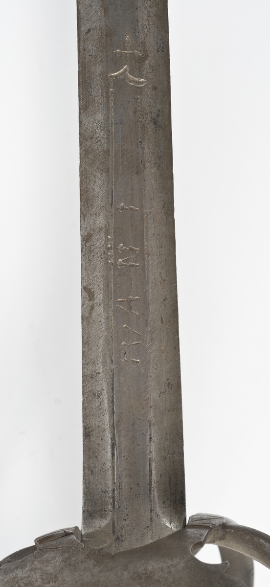 Plommeformet  8 kantet knapp. Tregrep.. Asymmetrisk feste med flate parerbøyler.  S formede parerstenger..Fremre parerstang går over i håndbøylen.Oppbøyet skjellformet parerplate på innsiden.
Tveegget, rett klinge med 6 kantet tverrsnitt  Kort blodrand merket IVAN