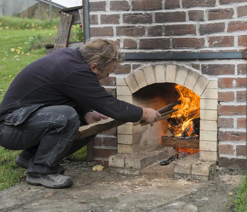 Mann i svarte klær sitter på huk og putter mer ved inn i flammene i den teglsteinsmurte kalkovnen.