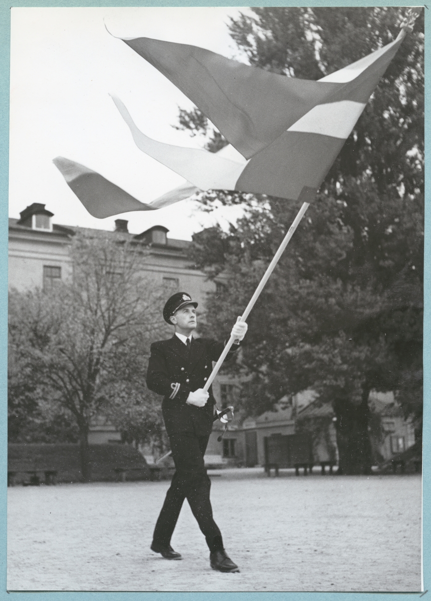 Bilder från Krigsmans erindran den 13/10 1951. En uniformsklädd löjtnant står med en tretungad flagga i händerna på bataljon Sparres kaserngård. I bakgrunden syns kasern Sparre som delvis skyms av träd.