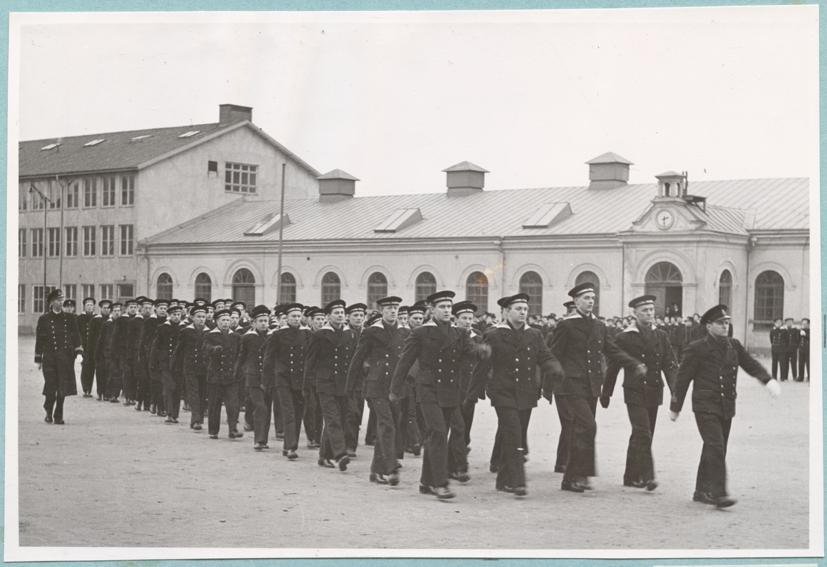 En grupp med flottister och militärer ur flottan marscherar över bataljon sparres kaserngård. I bakgrunden syns Sparres excercishus.