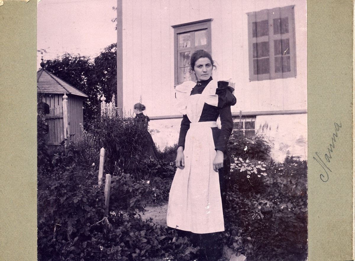 Kvinne står i hage foran hvit trebygning. Mørk kjole med hvitt forkle. Nanna Pedersen. Husveggen bak henne har et påmalt vindu. (Falsk vindu). En kvinne til skimtes bak noen stauder i hagen.