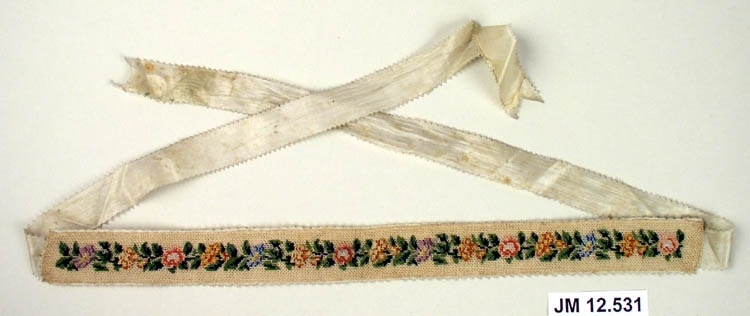 Broderat strumpeband, brodyren är fäst på sidenband. Blomstergirlang med blommor i olika färger, troligen använt och broderat av bruden själv. Paret till bandet saknas.