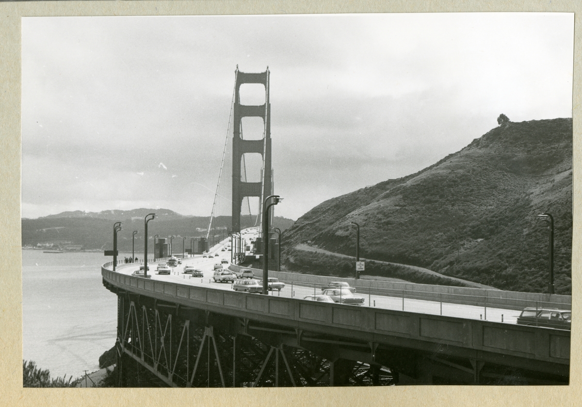 Bilden föreställer ett landskap med en stor vägbro, vilken det kör bilar över. Bilden är tagen under minfartyget Älvsnabbens långresa 1966-1967.