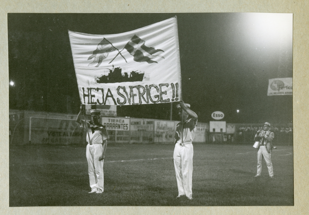 Bilden föreställer två uniformsklädda besättningsmän, vilka står på en fotbollsplan och håller i en banderoll med texten "HEJA SVERIGE". Minfartyget Älvsnabben och två svenska flaggor har även avbildats på banderollen. Bilden är tagen i Puntarenas i samband med Älvsnabbens långresa 1966-1967.