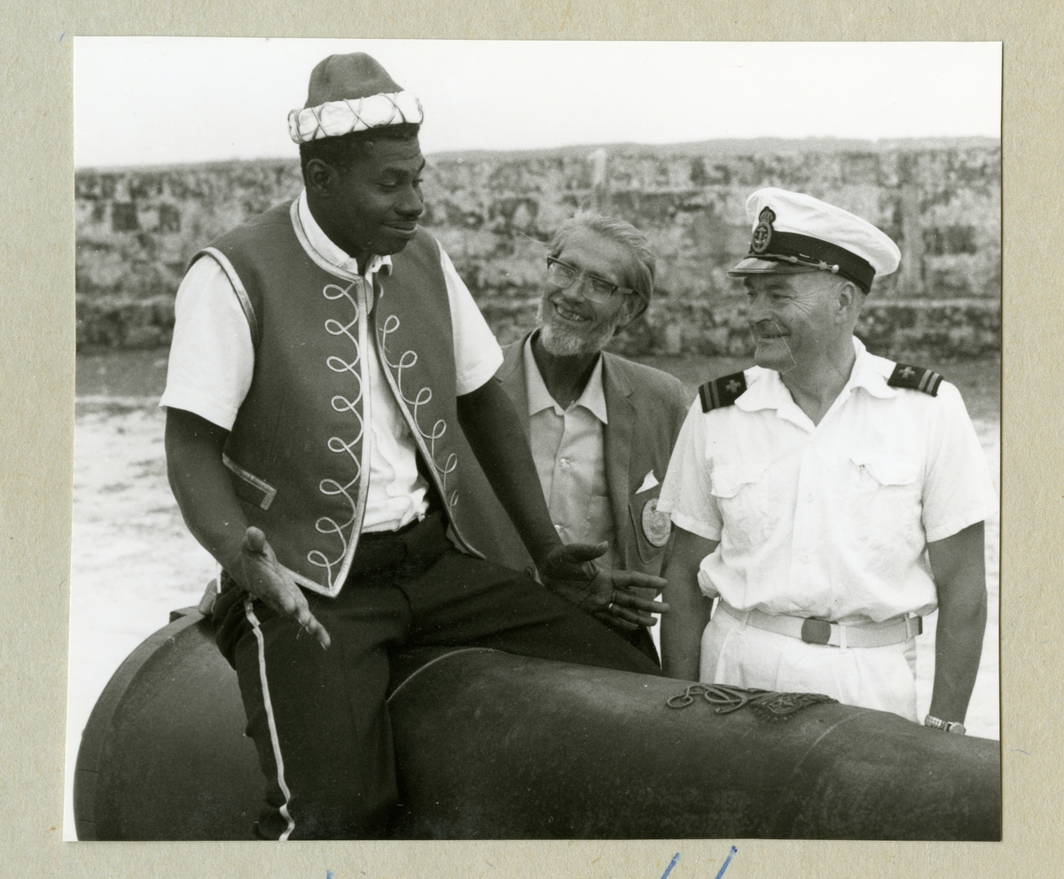Bilden föreställer tre män vid en kanon. En är klädd i vit uniform, en i kavaj och den sista som sitter på kanonen är klädd i väst. Bilden är tagen under minfartyget Älvsnabbens långresa 1966-1967.