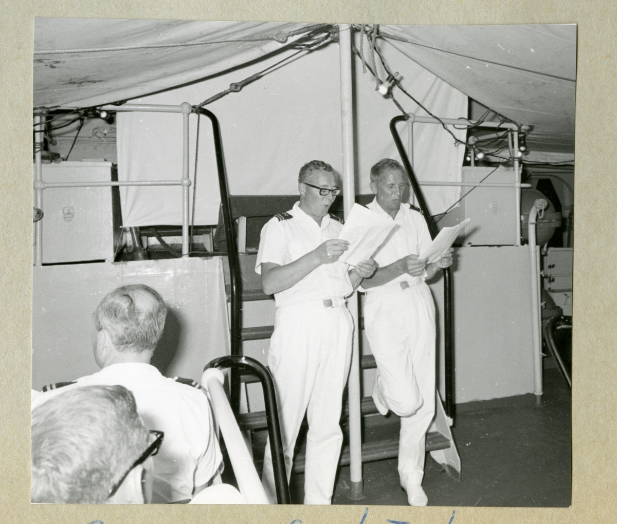 Bilden föreställer två uniformsklädda besättningsmän som läser i papper och spexar inför publik på minfartyget Älvsnabbens däck. Bilden är tagen under fartygets långresa 1966-1967.