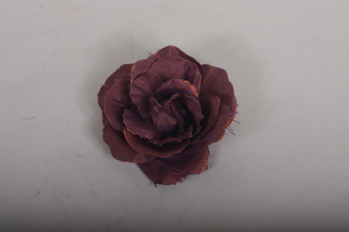 Forma som ei stor rose. På baksida er roseblada festa til eit stoffstykke forma som eit svart roseblad. På denne er også sjølve nåla festa.