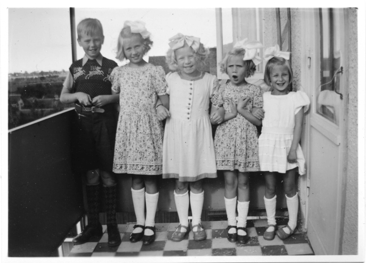 En pojke och fyra flickor med i klänning och stora rosetter i håret på balkong vid Brahegatan 59. Från vänster: Lennart Kjellström, Mona Kjellström, Birgitta Carlsson, IngBritt Kjellström och Vivianne Ehrenberg. Lennart, Mona och Ingbritt var syskon.