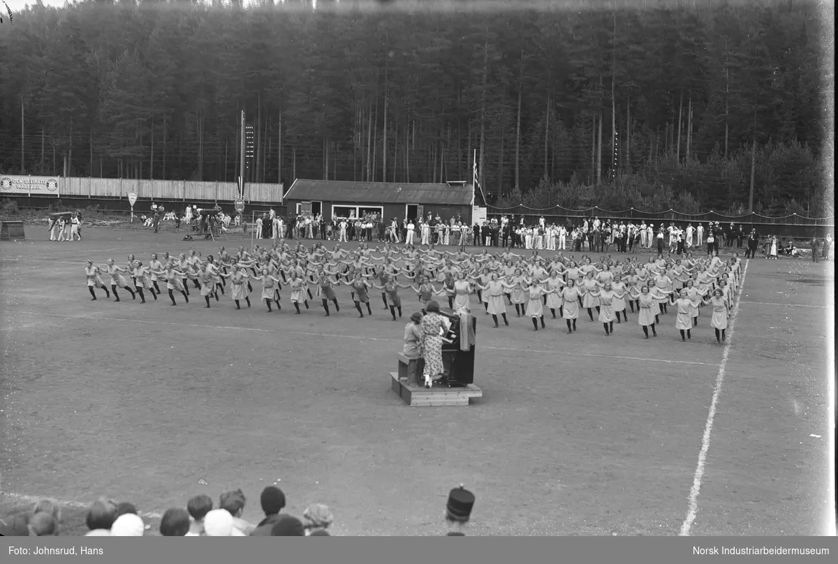 Turnstevene 1933. Turndamer har oppvisning, to kvinner ved piano på gresset. Tilskuere rundt stadion.