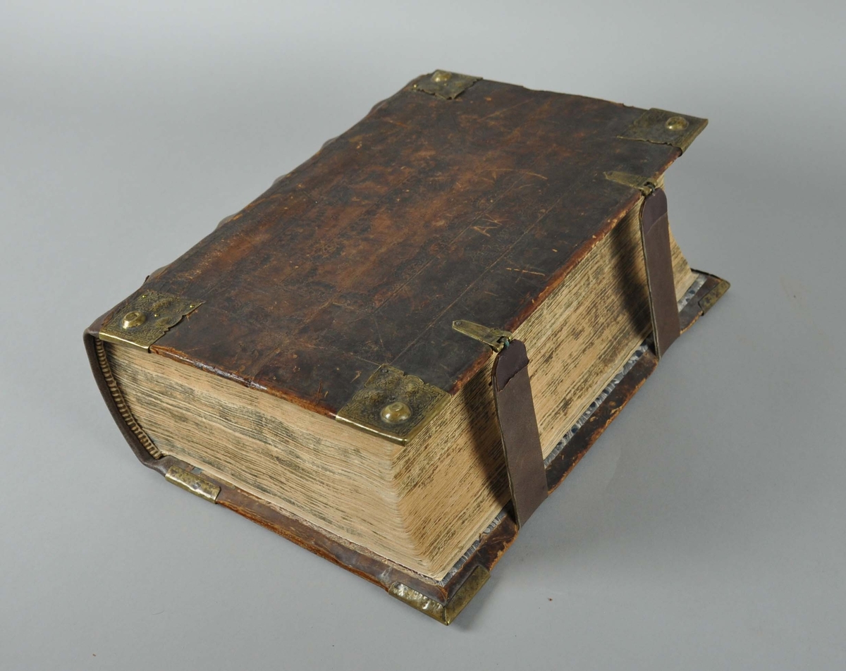 Bibel med brunt omslag av skinn. Bokomslaget har beslag av messing i hjørnene, og to festereimer av skinn.