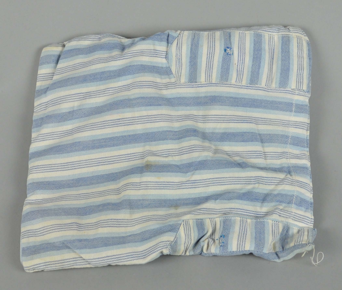 Madrass til dukkevogn av tekstil i hvitt og blått, med stripemønstre. Tekstilen er noe flekkete.
