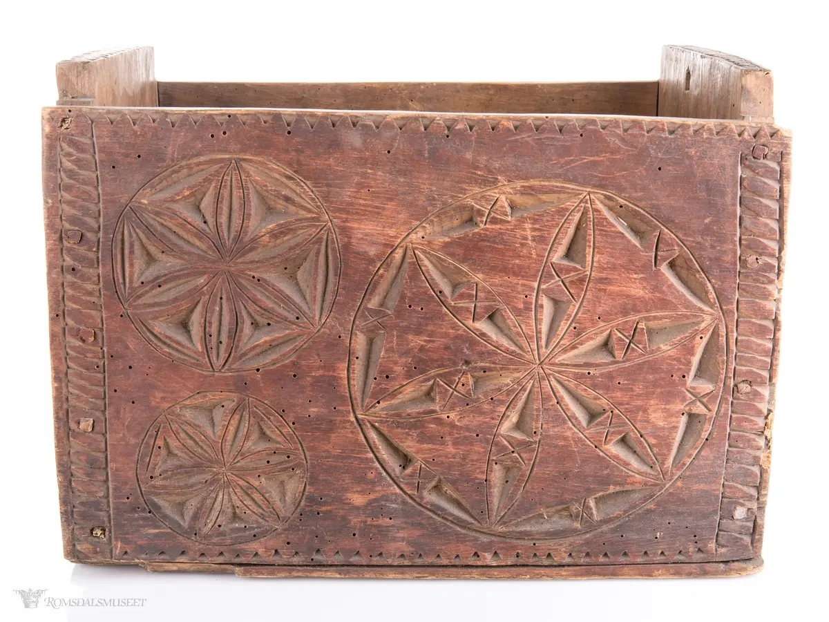 En stor og to små seksbladsroser i karveskurd på langsidene med skraverte dekorfelter og spetter rundt. På kortsidene er det skåret ut en stor seksbladsroser med rike dekorasjonsfelter bestående av spetter, streker diamantformer, sirkler med strek gjennom og solkors/kors. De største rosenen har kryss skåret inn på kronbladene.