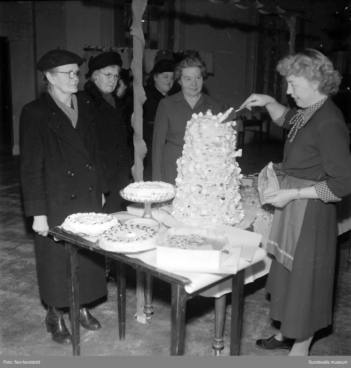 Hörselfrämjandets mässa på Församlingshuset. Kvinnor vid ett bord fullt av tårtor och andra godsaker, bland annat en imponerande spettekaka. En flicka och ett dockskåp.