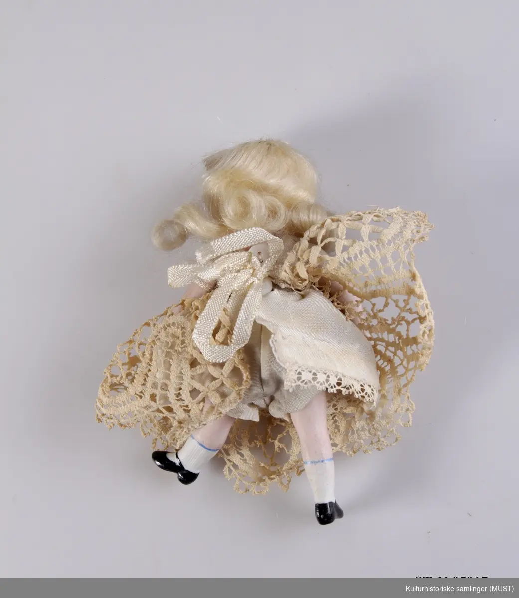 Porselendukke av jente med lyst langt hår og kjole med blondekant nederst. Hodet, armene og beina er bevegelige.